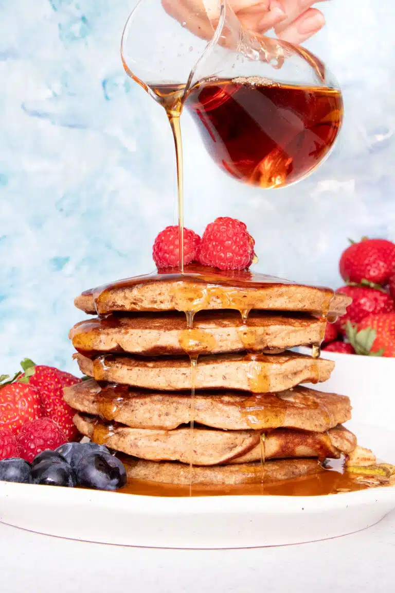 A large stack of Gluten Free Dairy Free pancakes is sitting on a white plate and being drizzled with maple syrup.