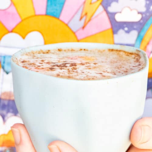 A white hand holds a small blue ceramic cup filled with a maca latte. The cup is held in front of a colourful background.