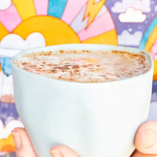 A white hand holds a small blue ceramic cup filled with a maca latte. The cup is held in front of a colourful background.