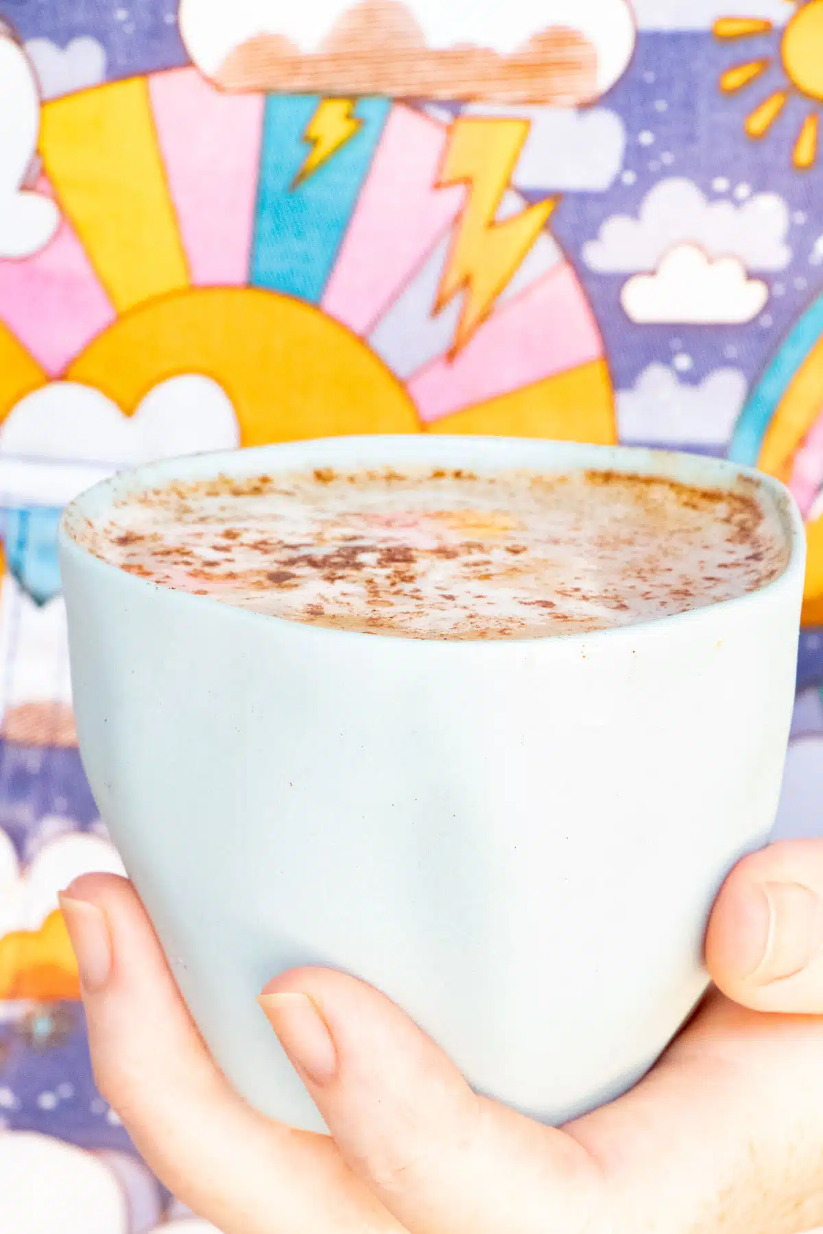 A white hand holds a small blue ceramic cup filled with a maca latte. The cup is held in front of a colourful background.