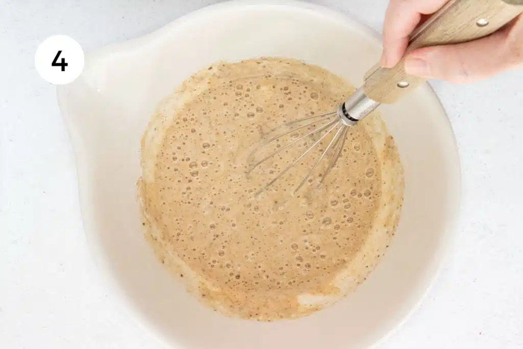 A white hand is gently whisking a gluten-free pancake batter in a large bowl.