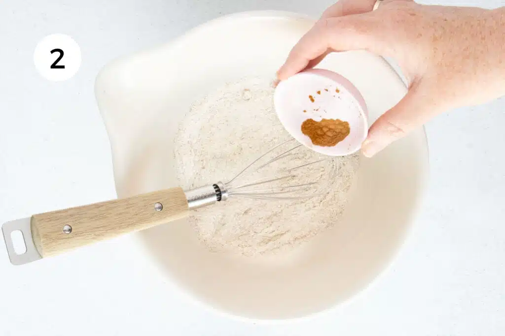 A white hand is tipping powdered cinnamon into a bowl filled with buckwheat flour and hazelnut meal.