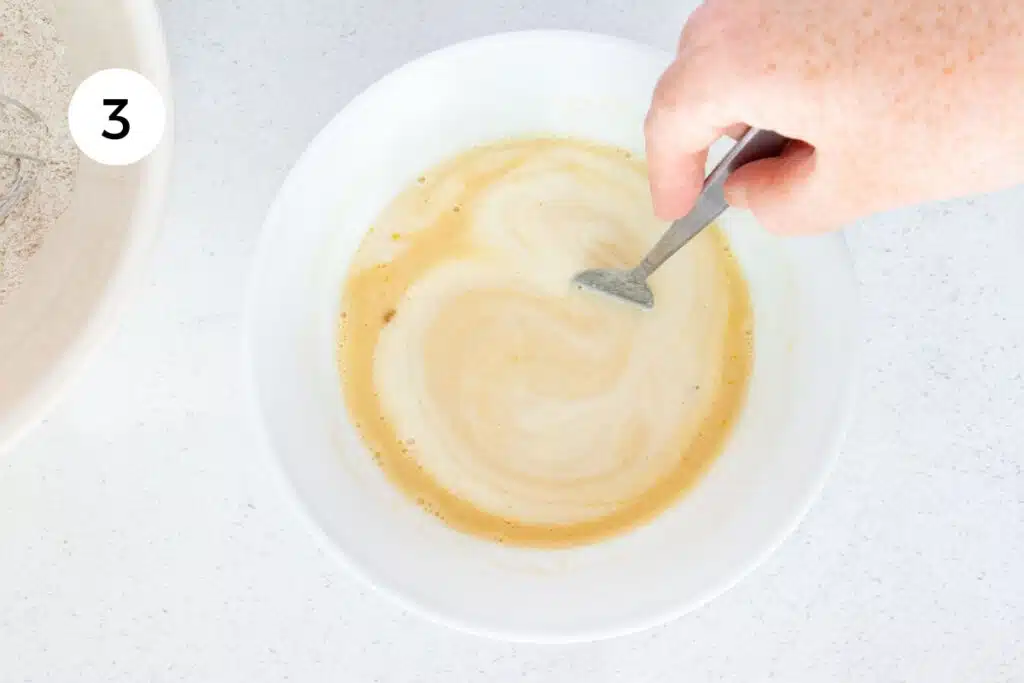 A white hand is stirring together dairy-free buttermilk, maple syrup and vanilla in a medium-sized bowl.