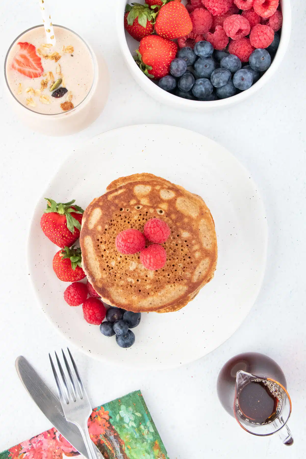 Looking down over a stack of pancakes on a plate set beside a smoothie and a small jug of maple syrup.