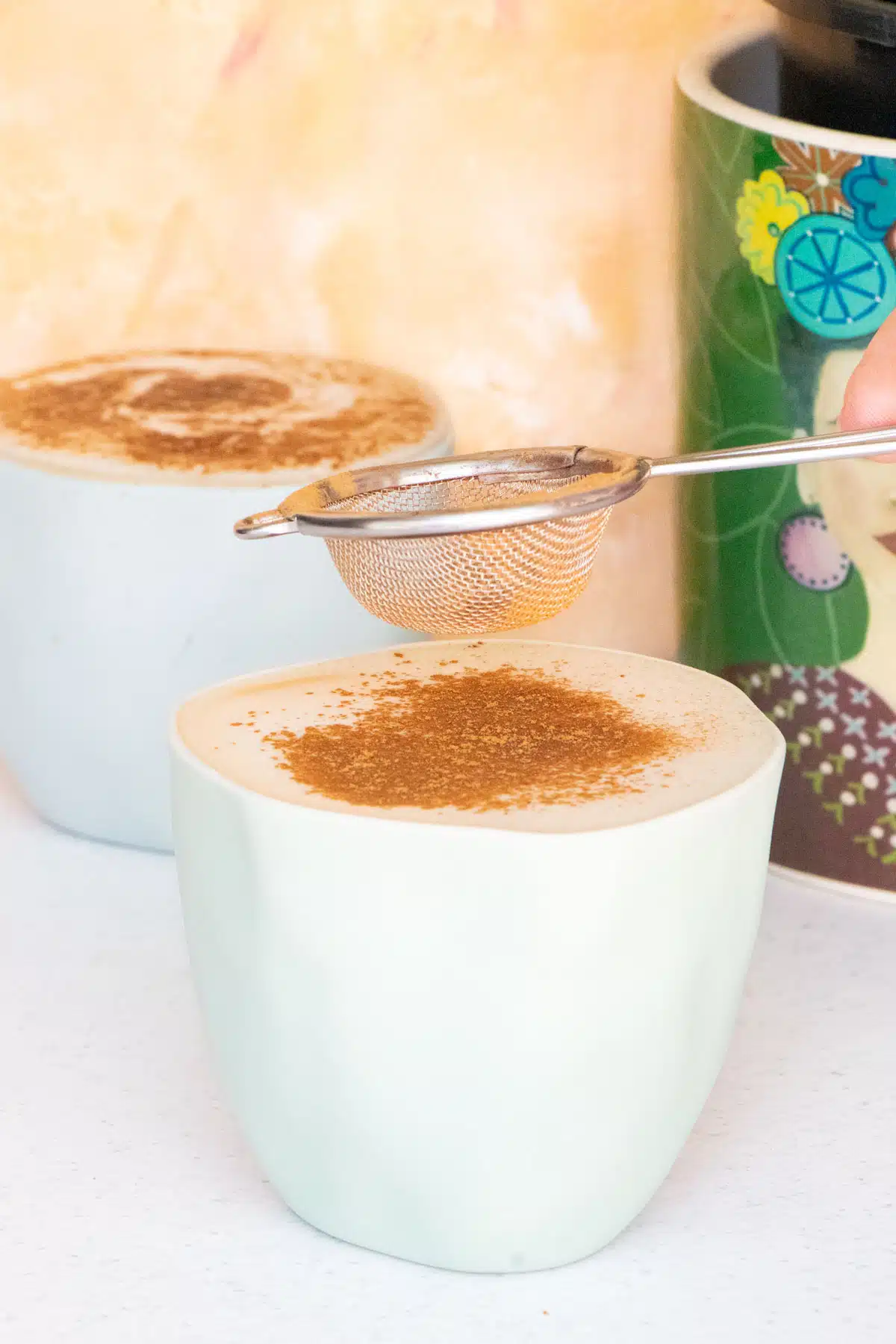 Cinnamon is being sifted through a very small sieve held above a blue ceramic coffee cup.