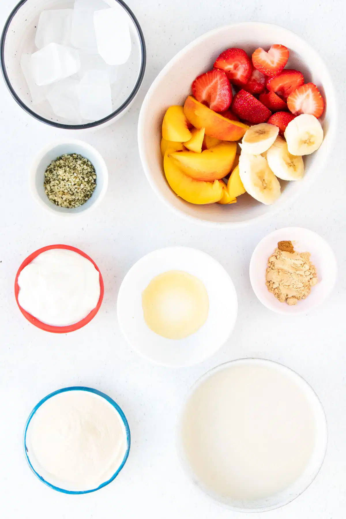 All the ingredients needed to make a peach and strawberry smoothie are laid out across a table in small bowls.