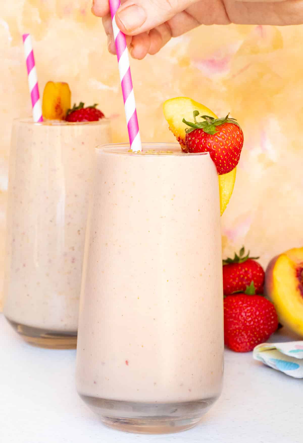 Two glasses filled with strawberry and peach smoothie decorated with a slice of peach and strawberry on the rim.