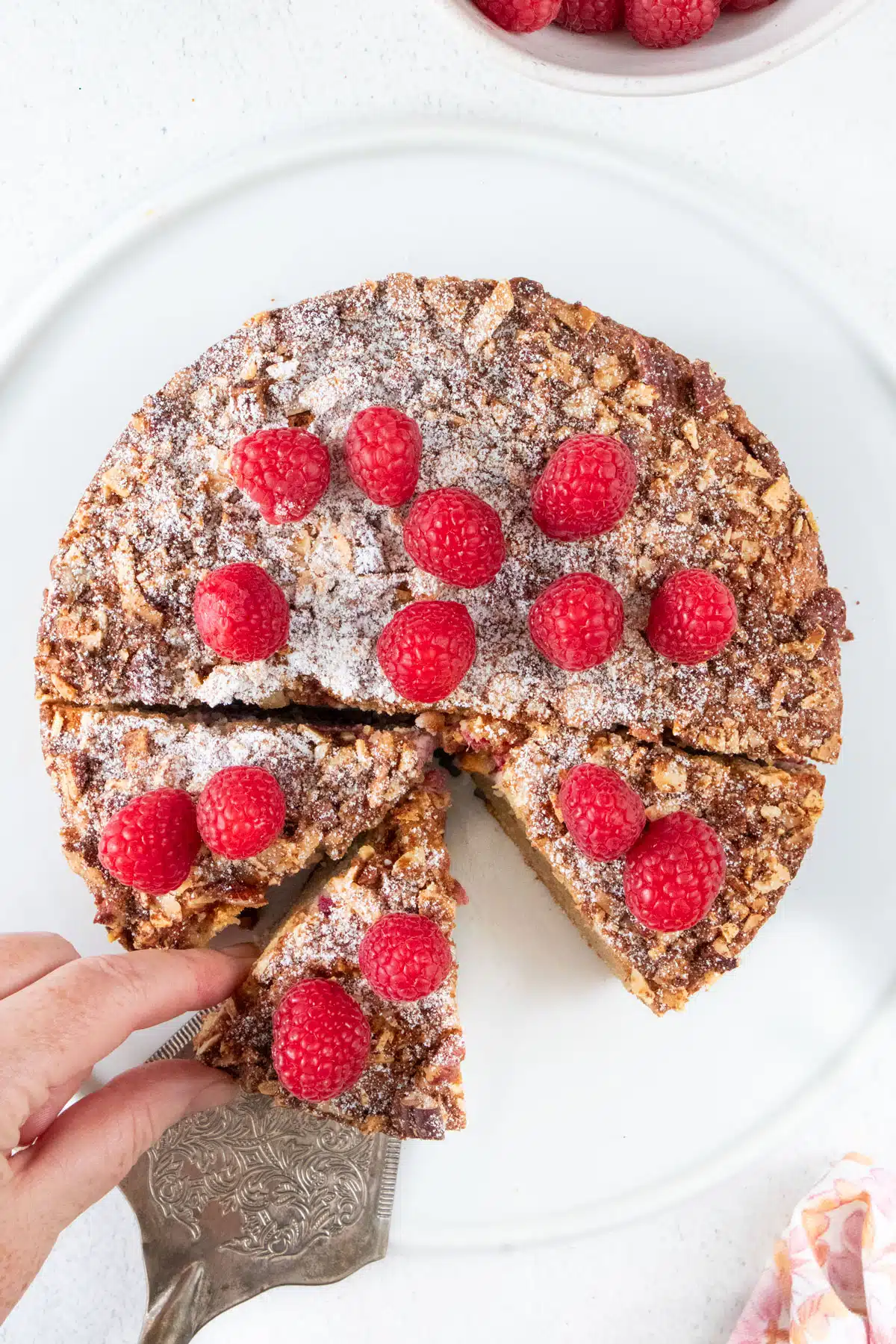 A white hand is using a cake server to take a piece of almond and raspberry cake from a white platter.