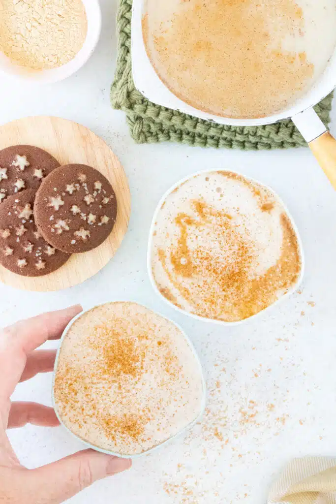 A white hand is picking up one of two cups of maca latte on a table. They sit beside a saucepan full of the milky mix.