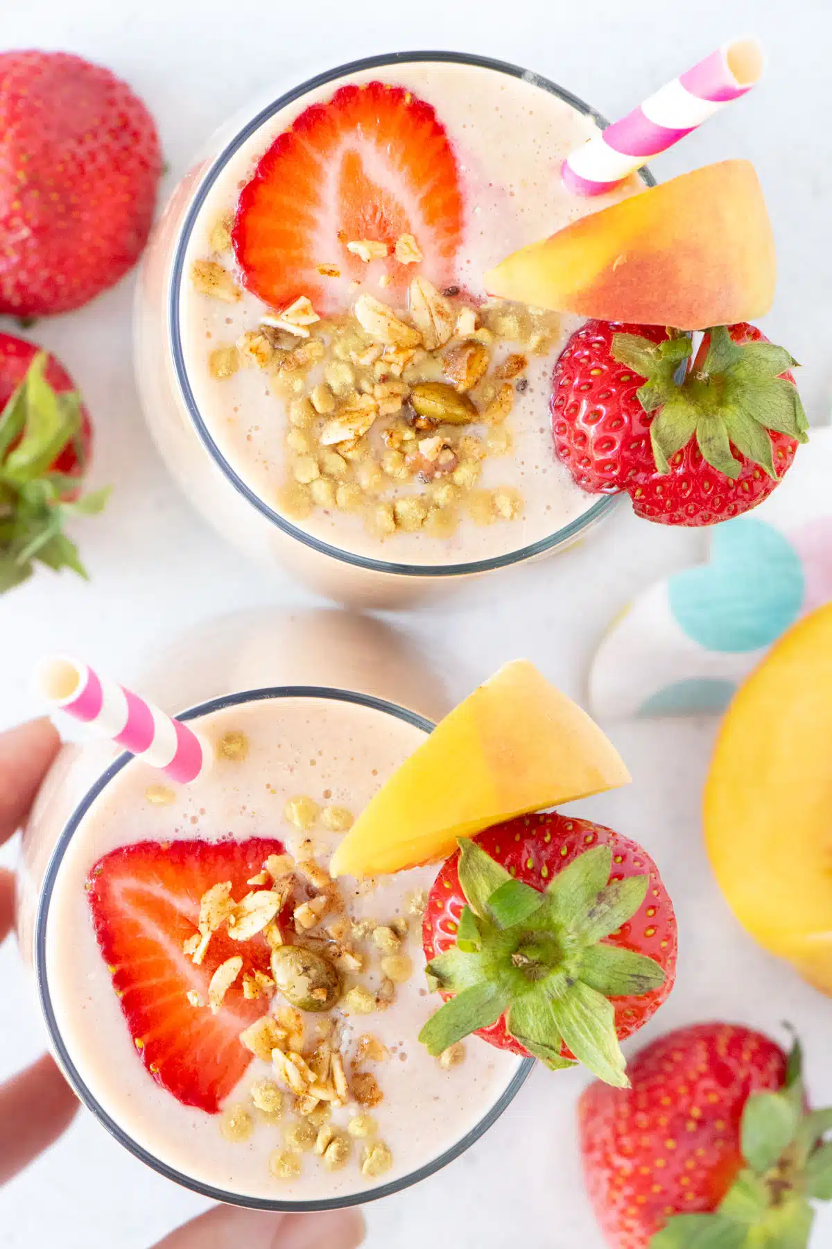 Two peach and strawberry smoothies are surrounded by strawberries and peach slices on a table.