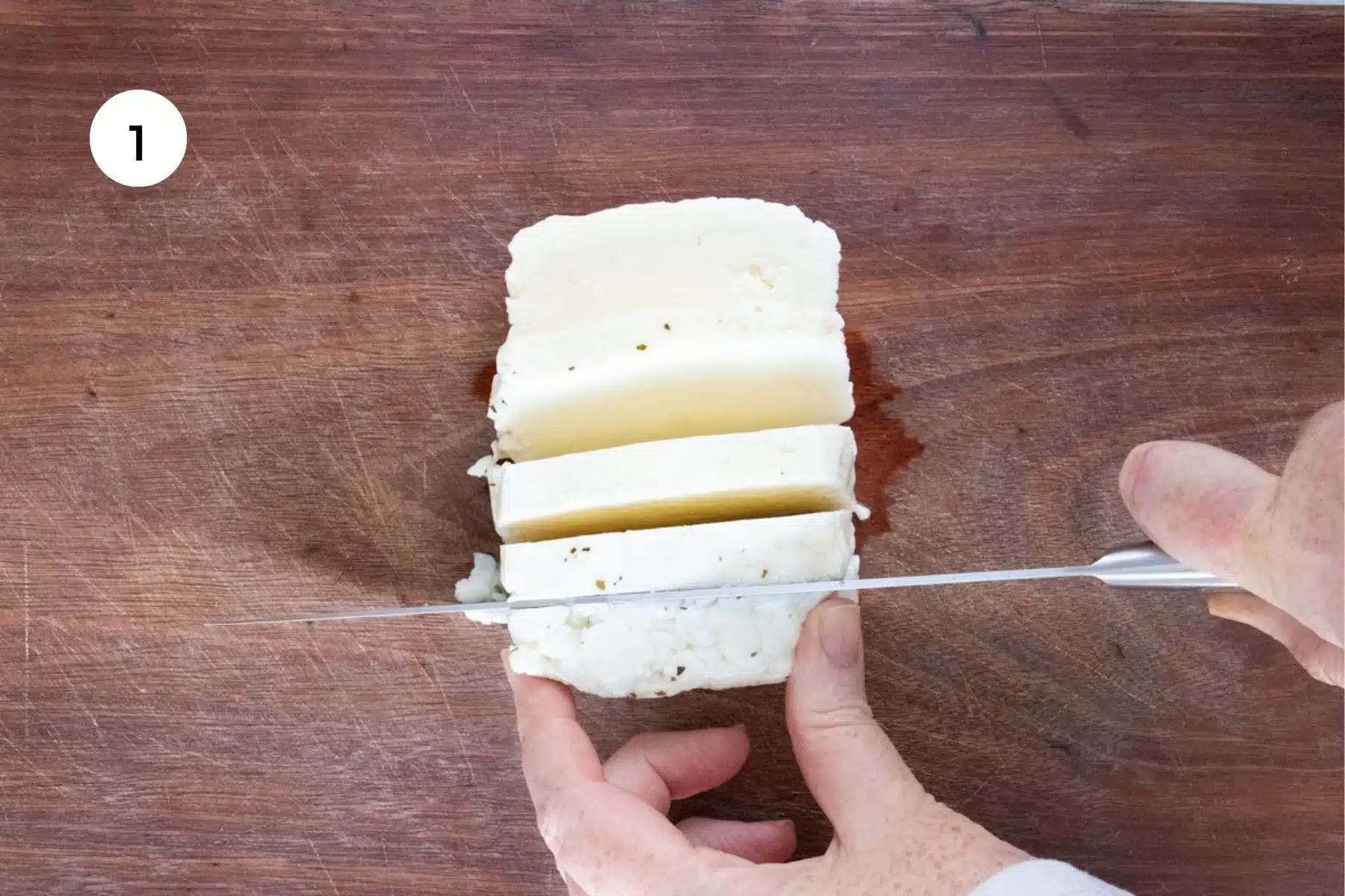 A white hand is slicing through a block of halloumi on a wooden chopping board with a large silver knife.