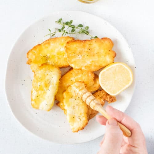 A white hand is drizzling honey over a plate of air fryer halloumi with a small wooden honey stirrer.