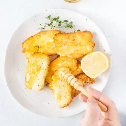 A white hand is drizzling honey over a plate of air fryer halloumi with a small wooden honey stirrer.