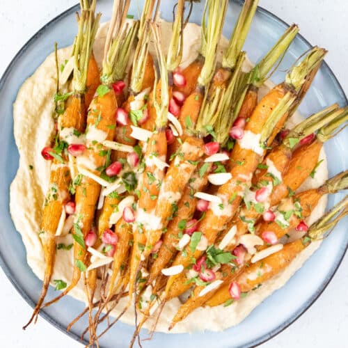 Maple roasted carrots are sitting on a bed of hummus on a large blue plate with a variety of toppings sprinkled over.