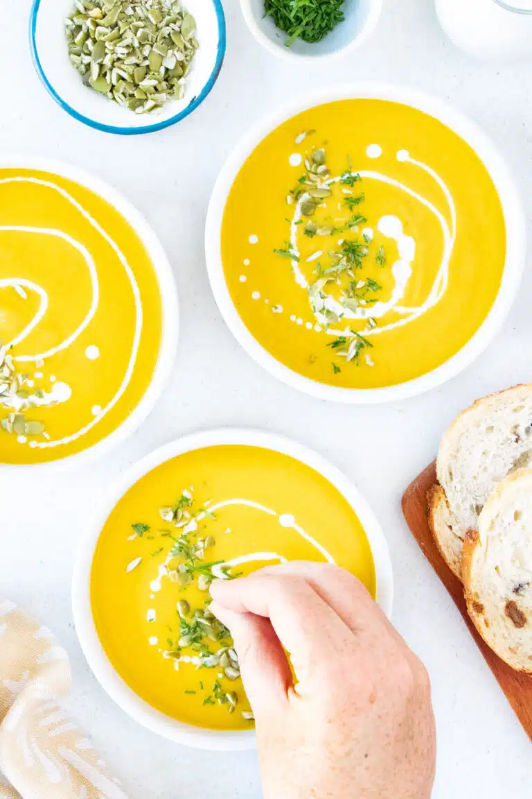 A white hand is sprinkled chopped green chives over a bowl of bright orange Instant Pot Pumpkin Soup.