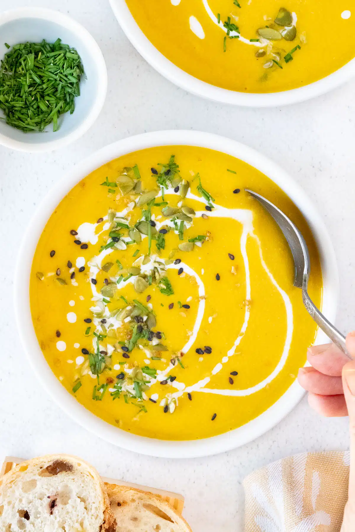 A white hand is using a silver spoon to take some bright orange pumpkin soup from a white bowl.