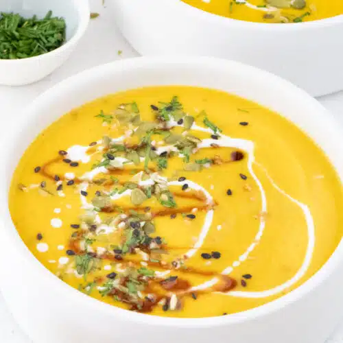 Two white bowls are filled with bright orange pumpkin soup and sprinkled with chopped seeds and chives.