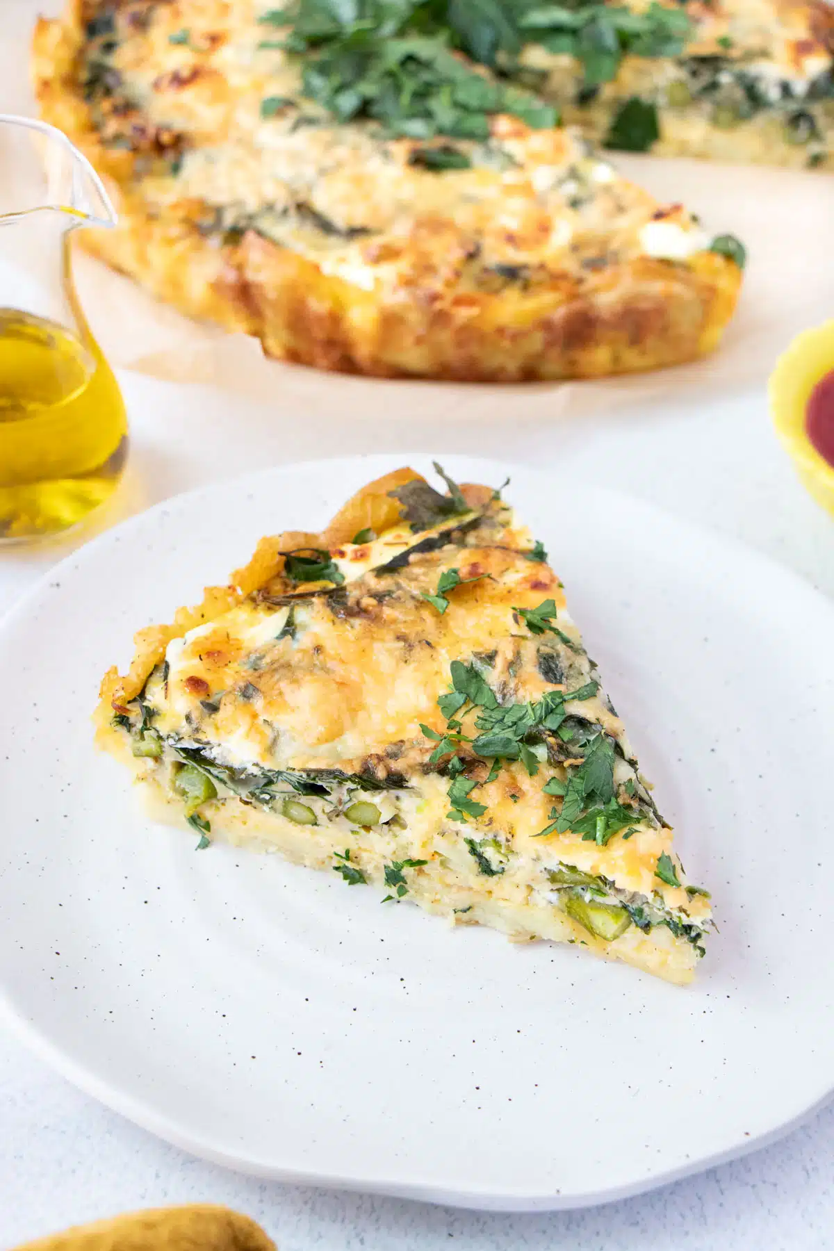 A slice of savory tart sits on a white plate with the full tart on a larger plate behind it.