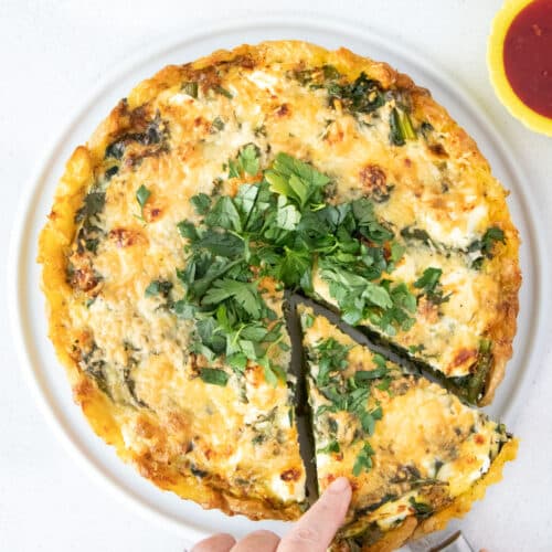 A white hand is sliding out a cut slice from a savory tart topped with chopped parsley.