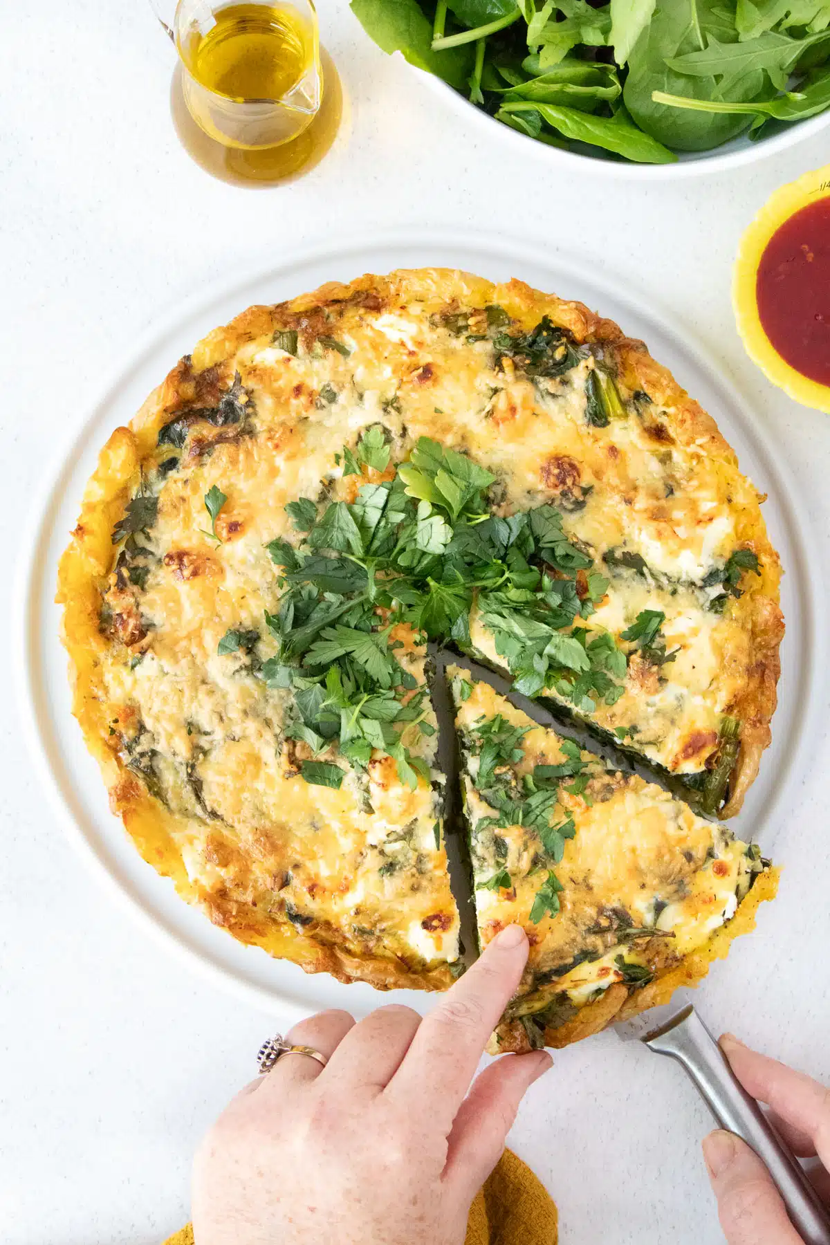 A white hand is sliding out a cut slice from a savory tart topped with chopped parsley.