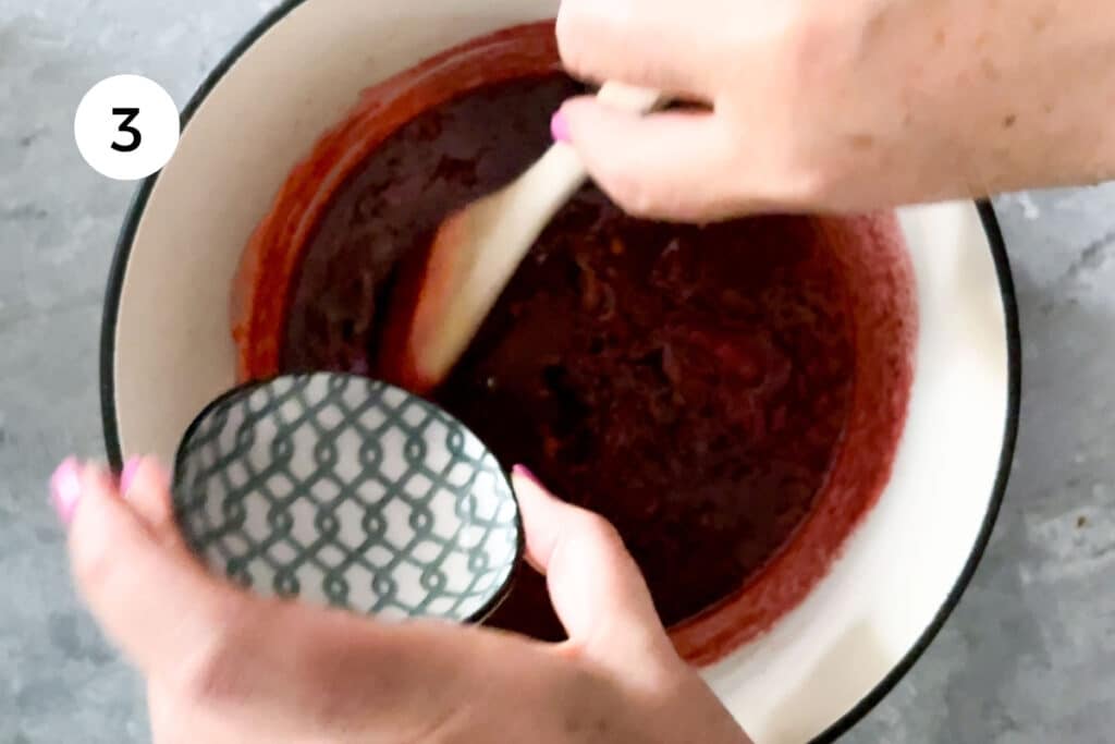 A white hand is pouring a small bowl of clear rose water into a pot of cooked strawberry jam.