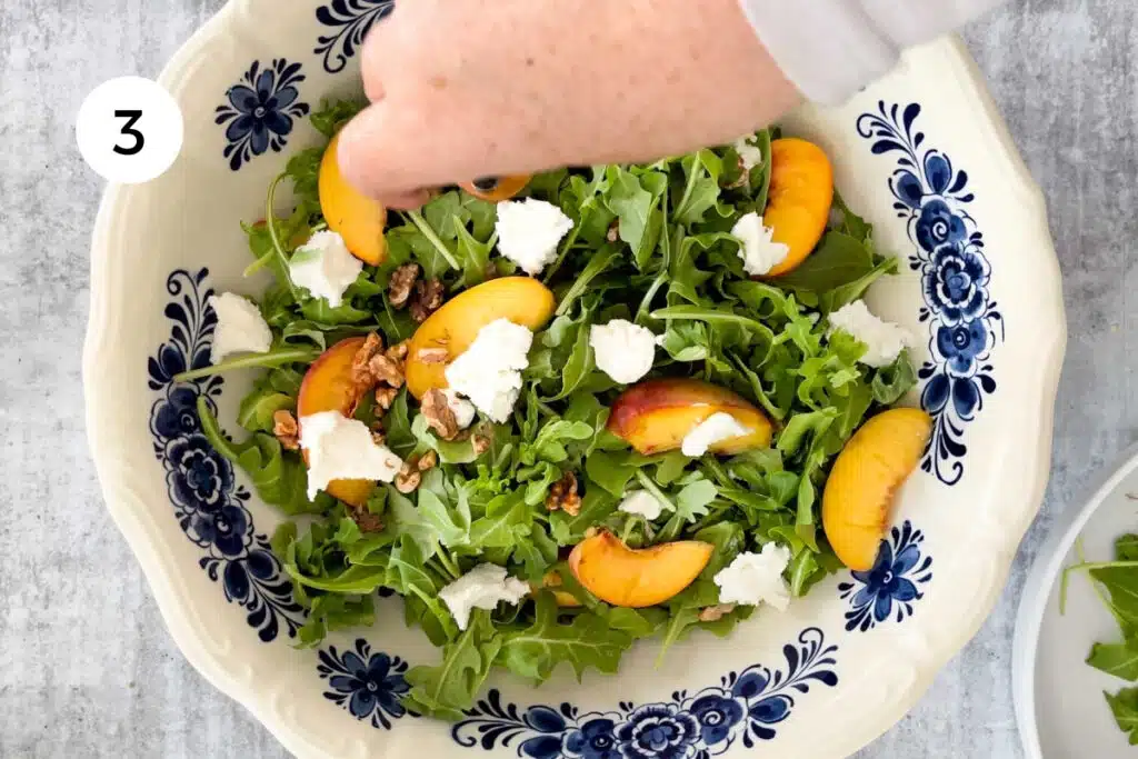 A white hand is crushing walnuts over a bowl of assembled peach arugula salad.