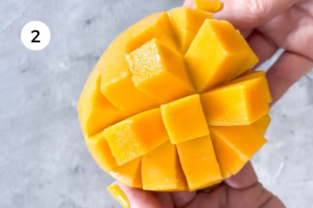 A mango cheek has been sliced in a crisscross pattern and two white hands are pulling it apart so the flesh pops up in cubes.