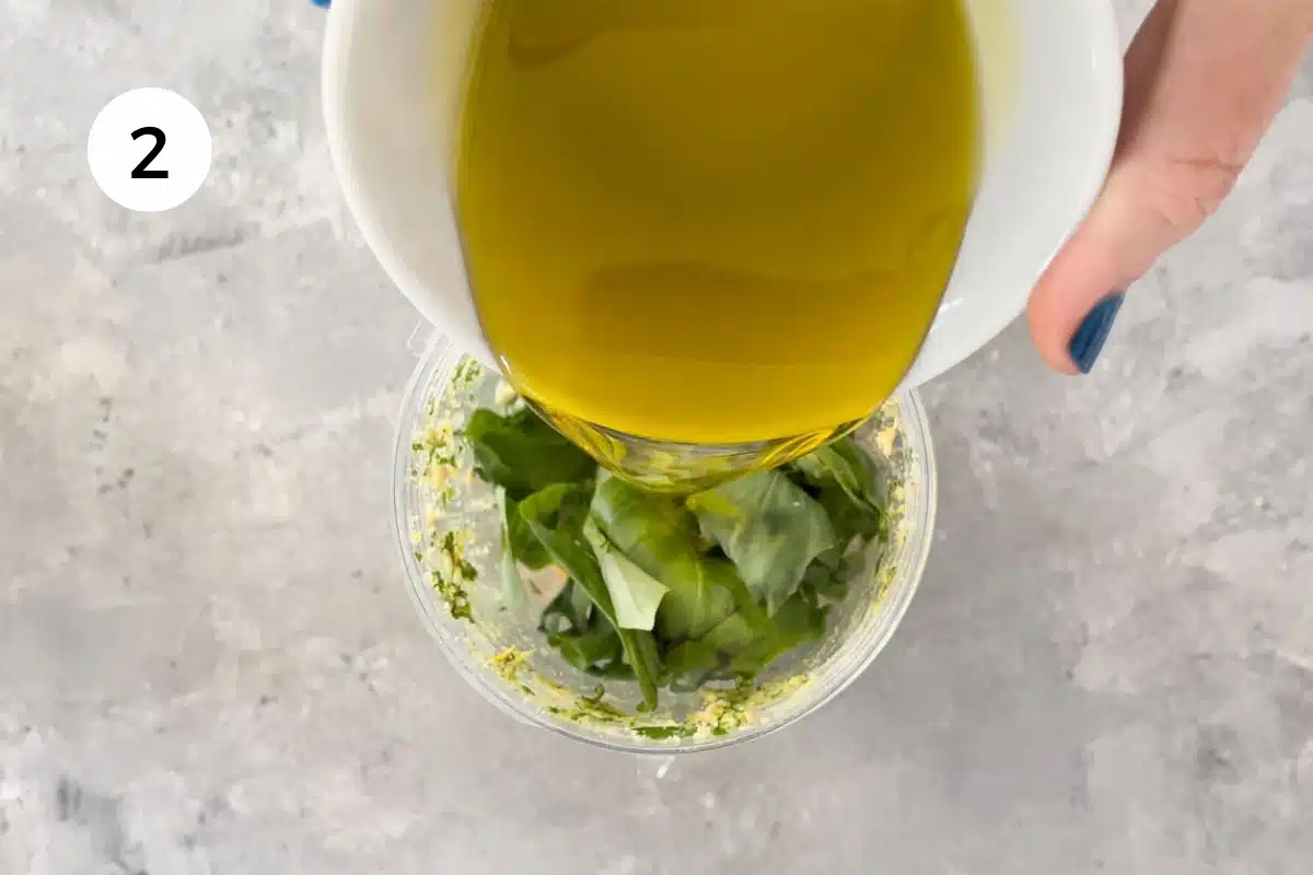 A white hand is pouring extra virgin olive oil into a nutribullet blender with other ingredients to make dairy free pesto.