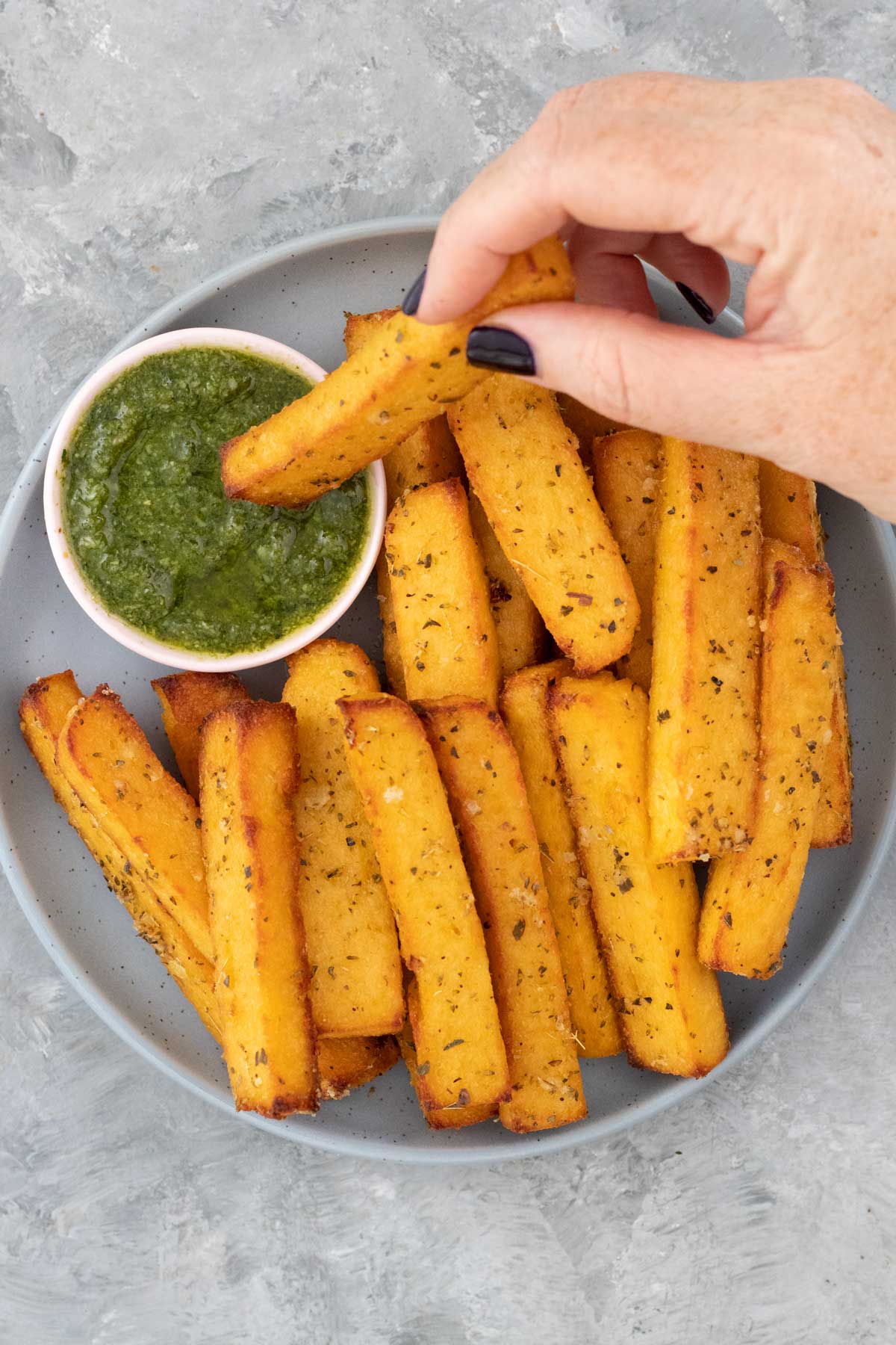 A white hand is dipping a baked polenta chip into a small bowl of green pesto.