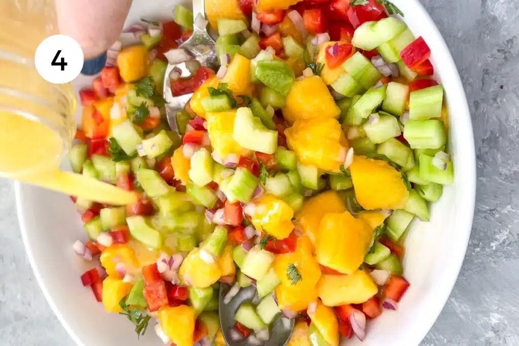 A white hand is pouring dressing over a chopped mango, cucumber, red capsicum/bell pepper, and red onion salad.