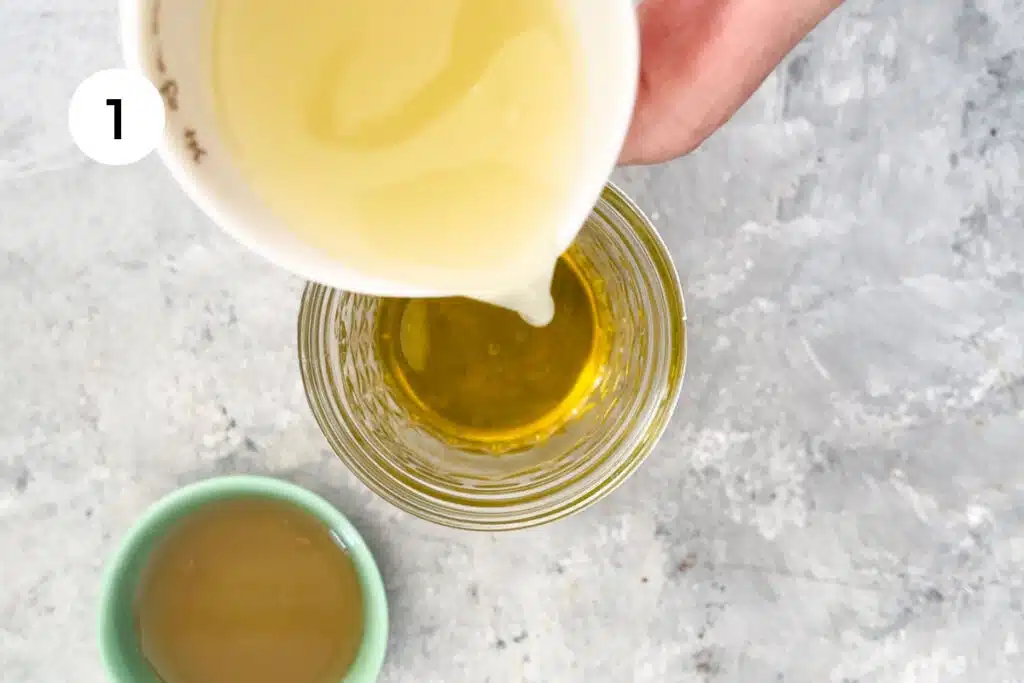 A white hand is pouring lemon juice into a small glass jar one third full with olive oil.