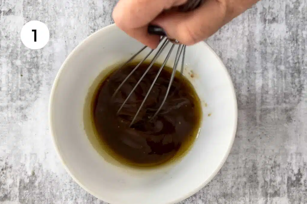 A white hand is mixing a salad dressing for the peach arugula (rocket) salad with a whisk in a white bowl.