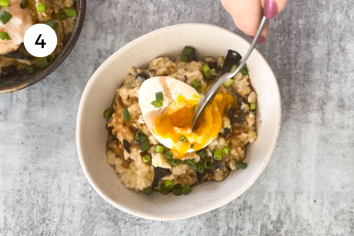 A white hand holding a silver spoon has pierced a poached egg on savoury oats.