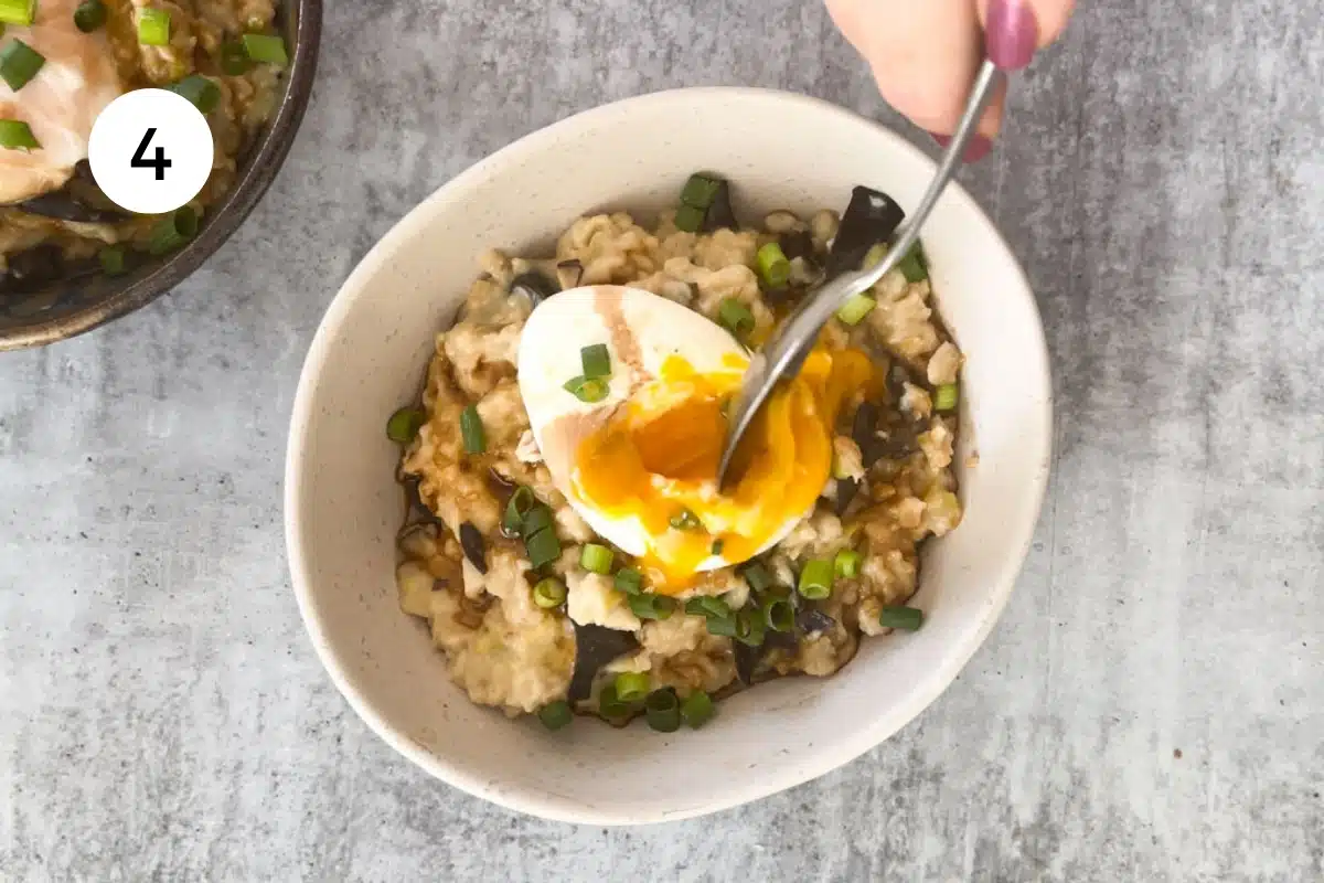 A white hand holding a silver spoon has pierced a poached egg on savoury oats.