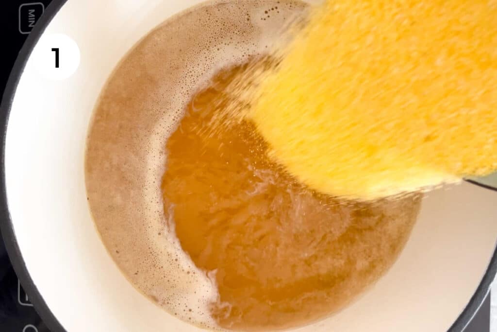 A cup of yellow polenta cornmeal is being poured into a pot of boiling chicken stock.