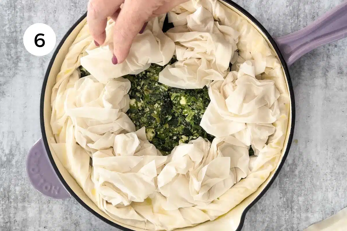 Two white hands are rolling up the overhanging edges of filo to create a crust around the edge of a skillet spanakopita.