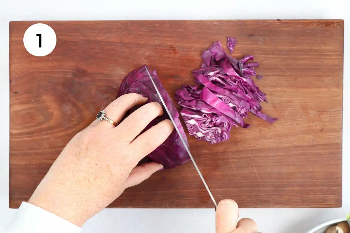 A white hand is holding a red cabbage on a wooden chopping board while the other hand slices it.