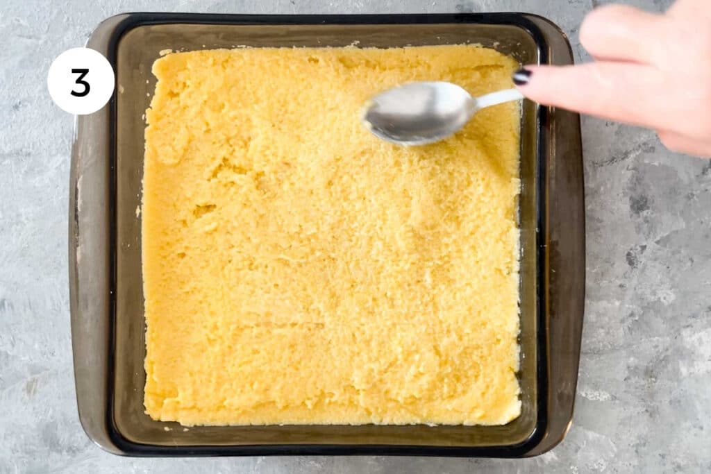 A brown glass baking dish has been filled with cooked polenta, which is being smoothed out with the back of a spoon.