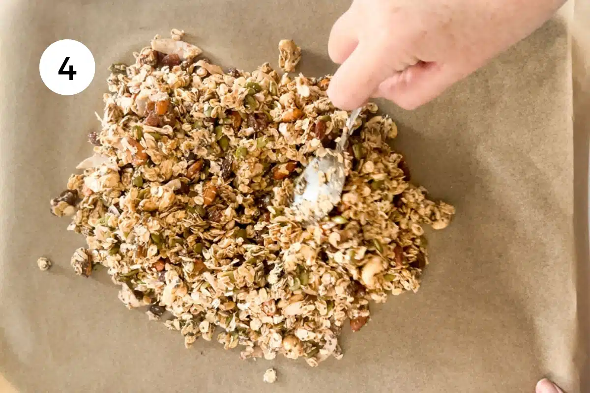 A white hand is using a spoon to spread the granola out on a baking tray lined with brown baking / parchment paper.
