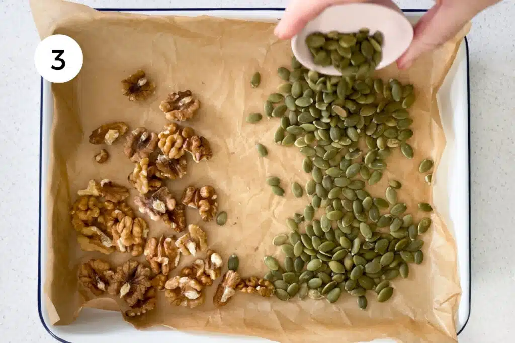 Nuts and seeds are being poured onto a baking tray lined with baking paper.