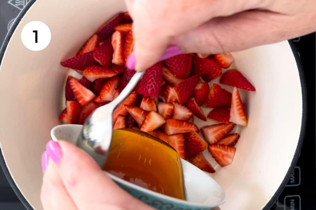A white hand is scraping honey from a small bowl with a teaspoon into a large pot of chopped strawberries.
