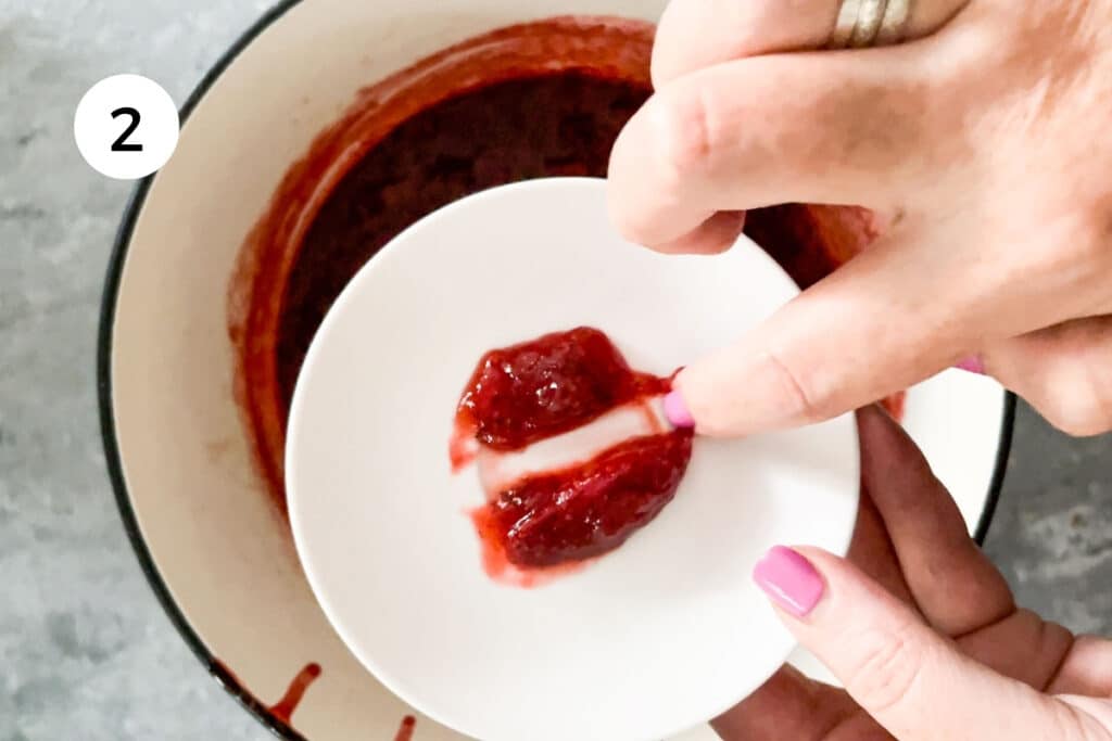 A white hand is running a finger through a big dollop of strawberry jam on a white saucer to check for doneness.