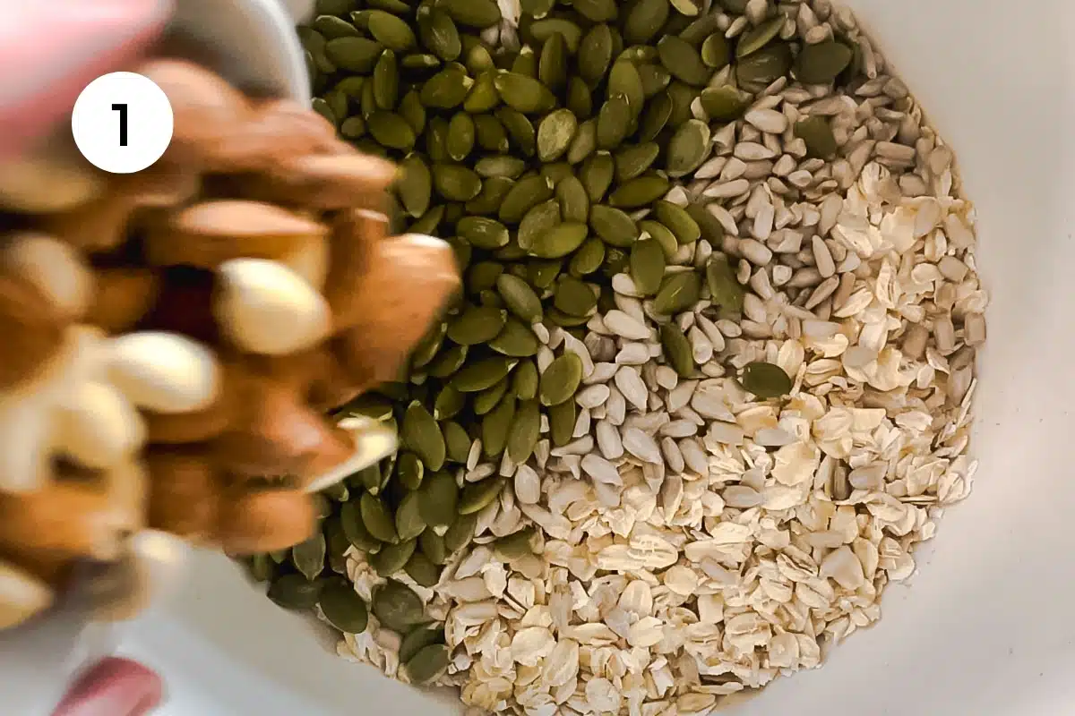 A white hand is tipping a cup of nuts into a large bowl filled with oats and seeds.