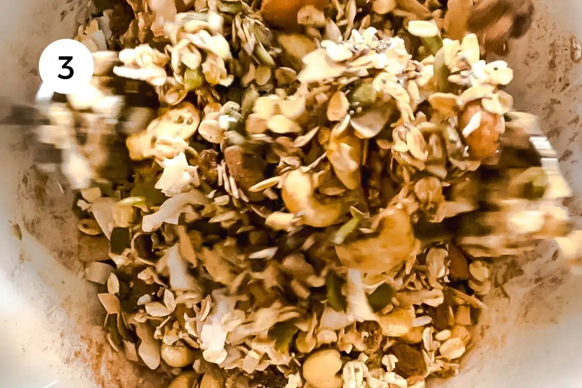 The granola mix is being tossed with two spoons in a large bowl.