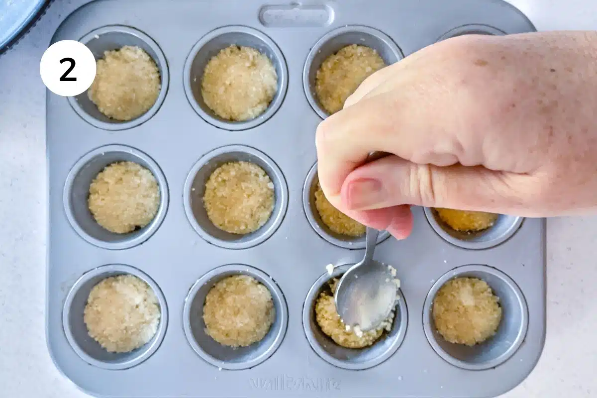 A white hand holding a spoon is pressing Twix cookie base into a mini muffin tin.