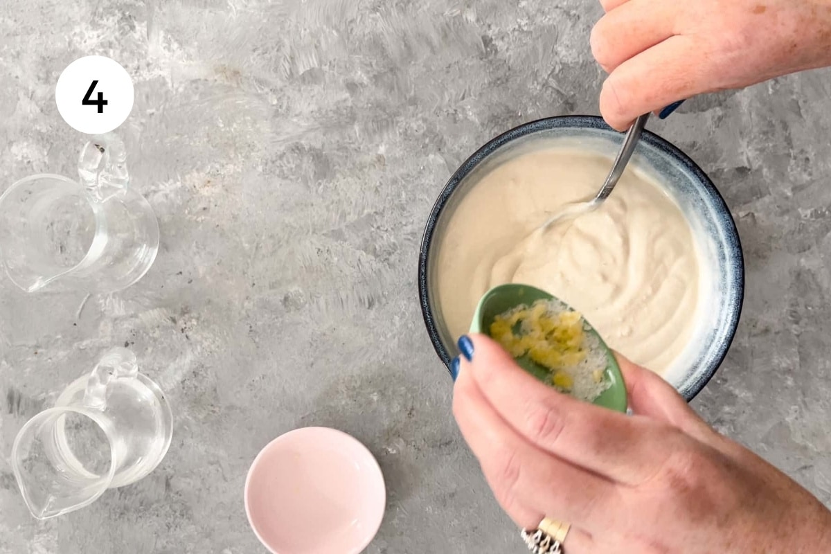 A small bowl of garlic and salt is being poured into a bigger bowl of lemon tahini sauce.