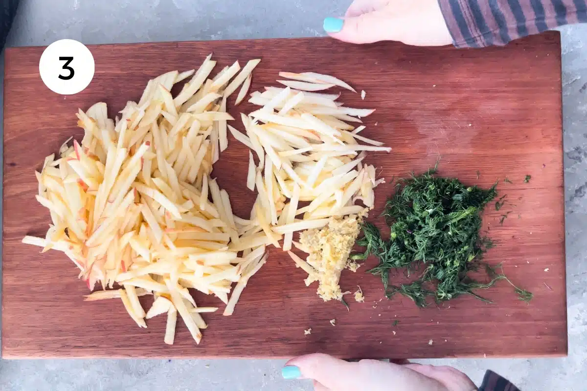 Grated apple, ginger and chopped fennel fronds are on a wooden chopping board being held by two white hands.