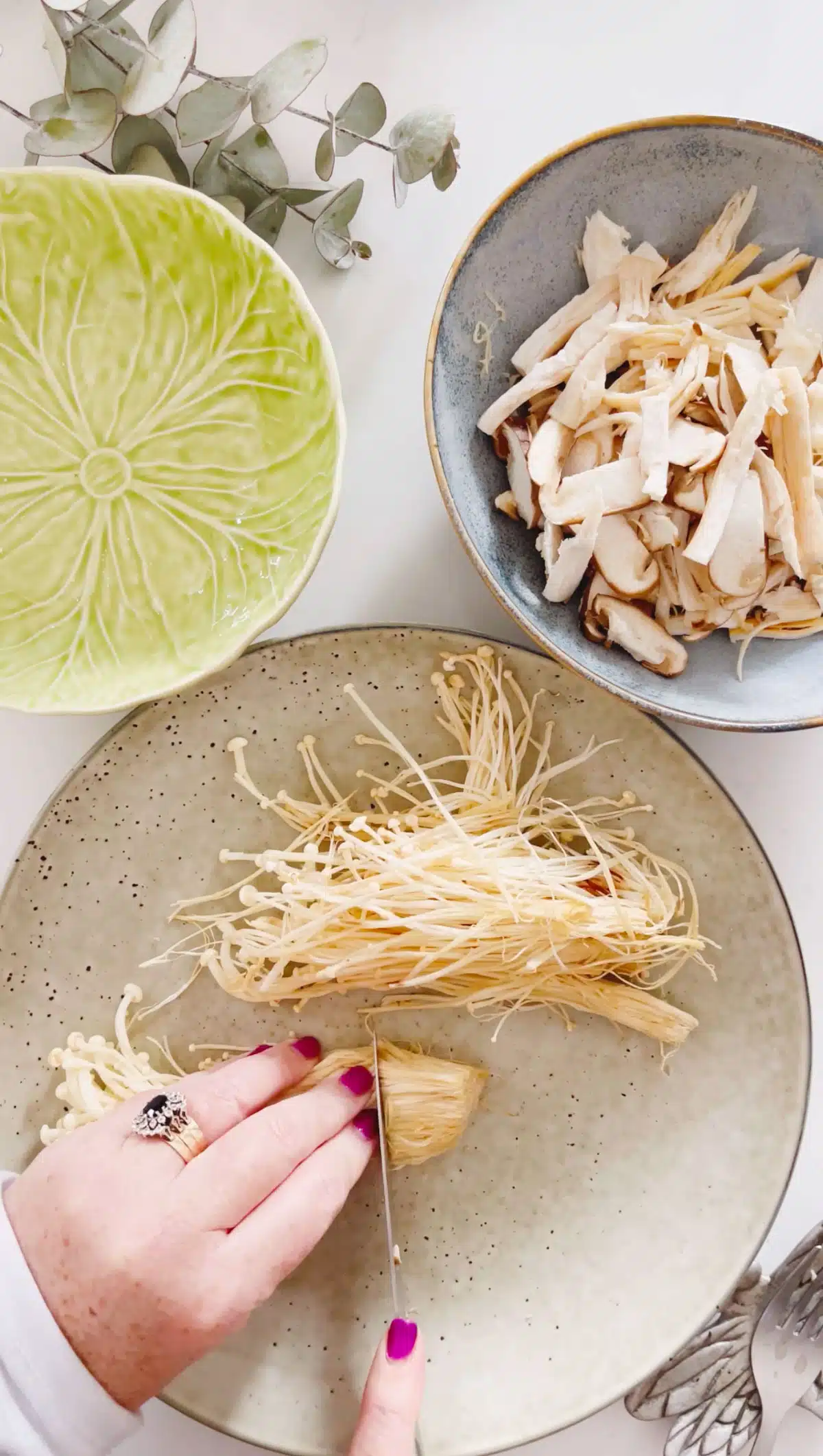 Two white hands a holding down a bunch of enoki mushrooms and cutting the ends off.