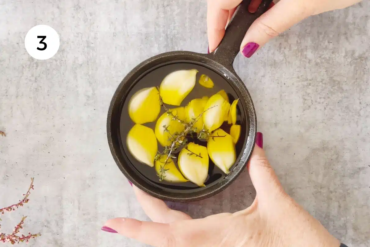 Two white hands are holding a small cast iron frying pan filled with garlic cloves, olive oil, and fresh thyme.