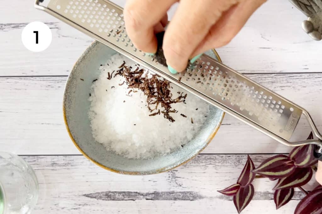 A white hand is using a microplane to grate fresh black truffle over a bowl of sea salt.